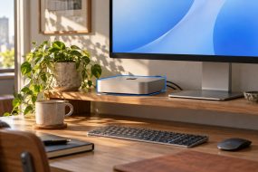 Compact desktop computer beside a monitor, keyboard and mouse on a desk.